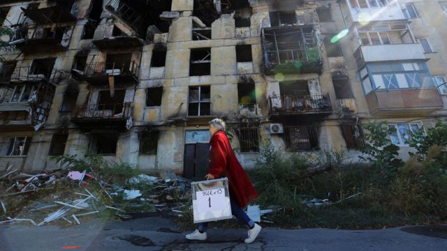 A woman carrying a clear portable ballot bag walks past a residential building severely damaged by an explosion and fire in Mariupol