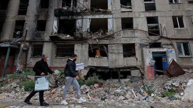 A man carrying a ballot bag and a woman walk past rubble and a building with severe damage in Mariupol