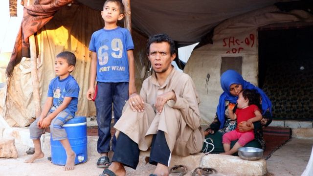 A man sitting on a stool with two children on his right, and a woman in blue headscarf holding a baby in the background