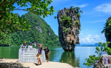 นักท่องเที่ยวยืนเซลฟี่กับป้ายเขาตาปู ด้านหลังมีวิวทิวทัศน์เขาตาปูตั้งตระหง่านอยู่กลางอ่าวพังงา