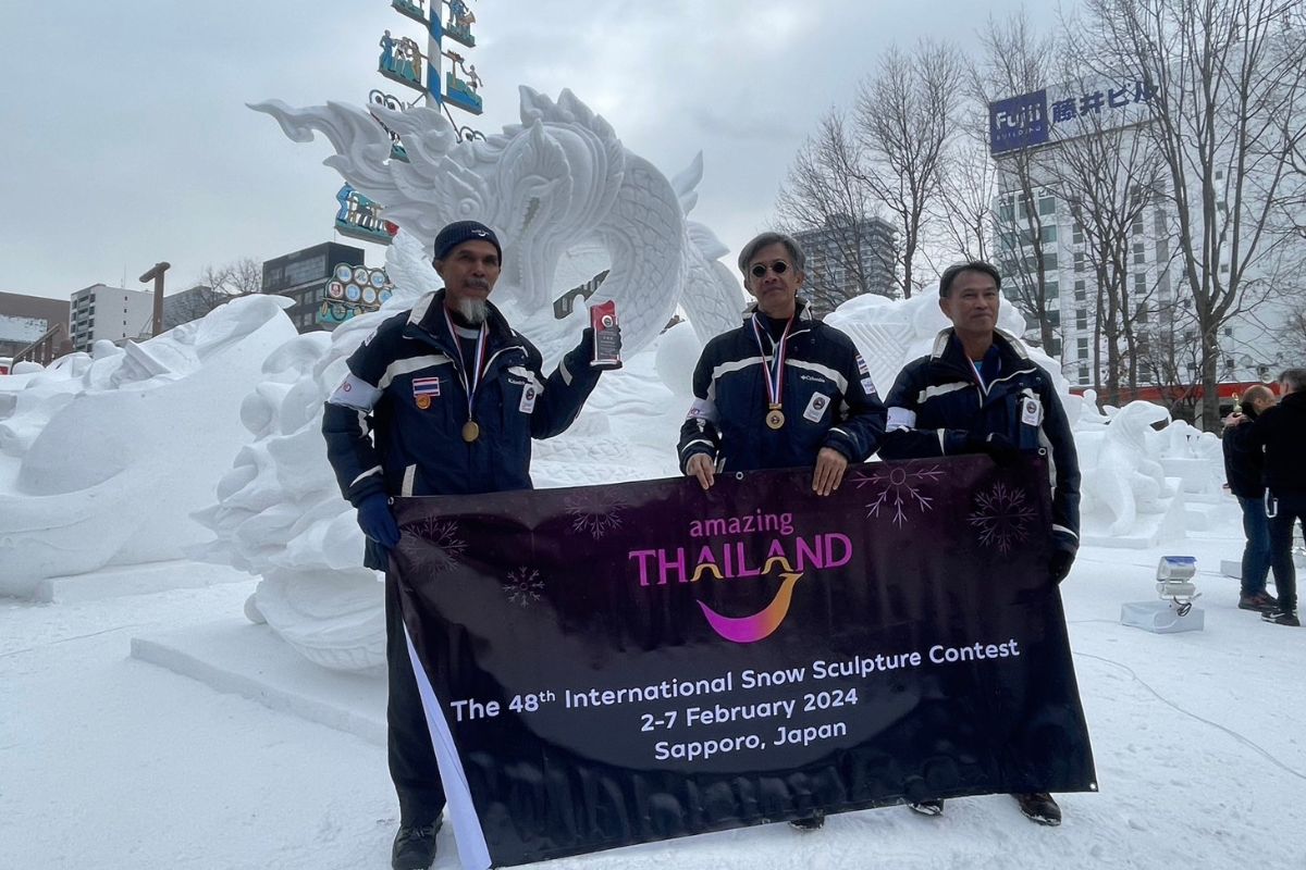 นักแกะสลักหิมะยืนถือถ้วยรางวัลด้านข้างผลงานแกะสลักหิมะรูปพญานาค ในบรรยากาศรอบข้างเต็มไปด้วยหิมะ