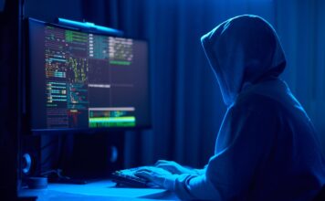 In a dimly lit room, a cloaked figure types code on a computer, hinting at a mysterious hacker